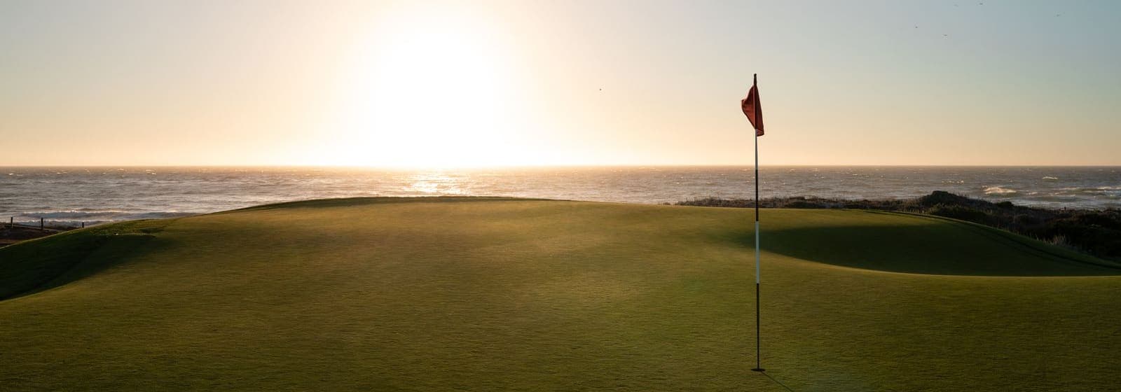 A golf green with a flag overlooking the Pacific Ocean at sunset in California