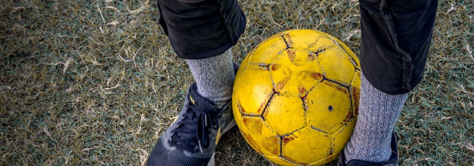 A pair of dark sneakers beside a worn yellow soccer ball on dry grass