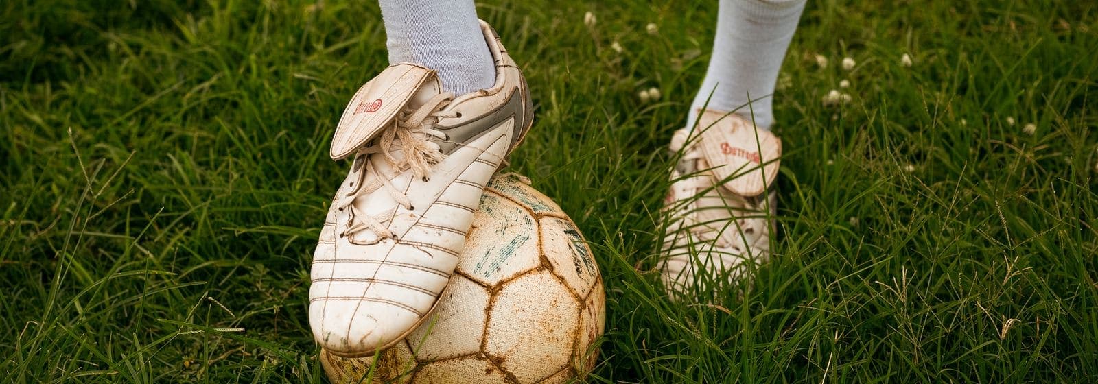 A player's cleat resting on a leather soccer ball in the grass