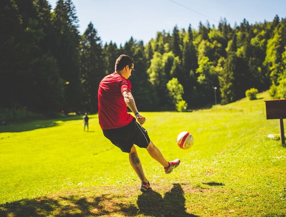 A man kicking a soccer ball across a sunny open field with tall trees behind him