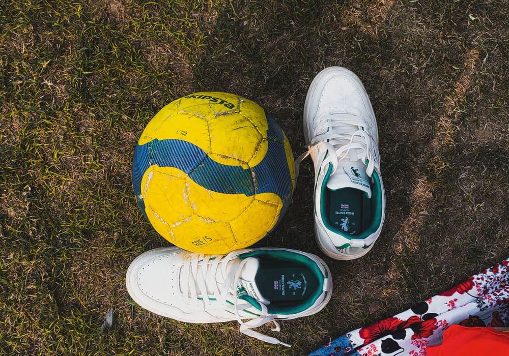A yellow soccer ball beside a pair of white sneakers laid out on the ground